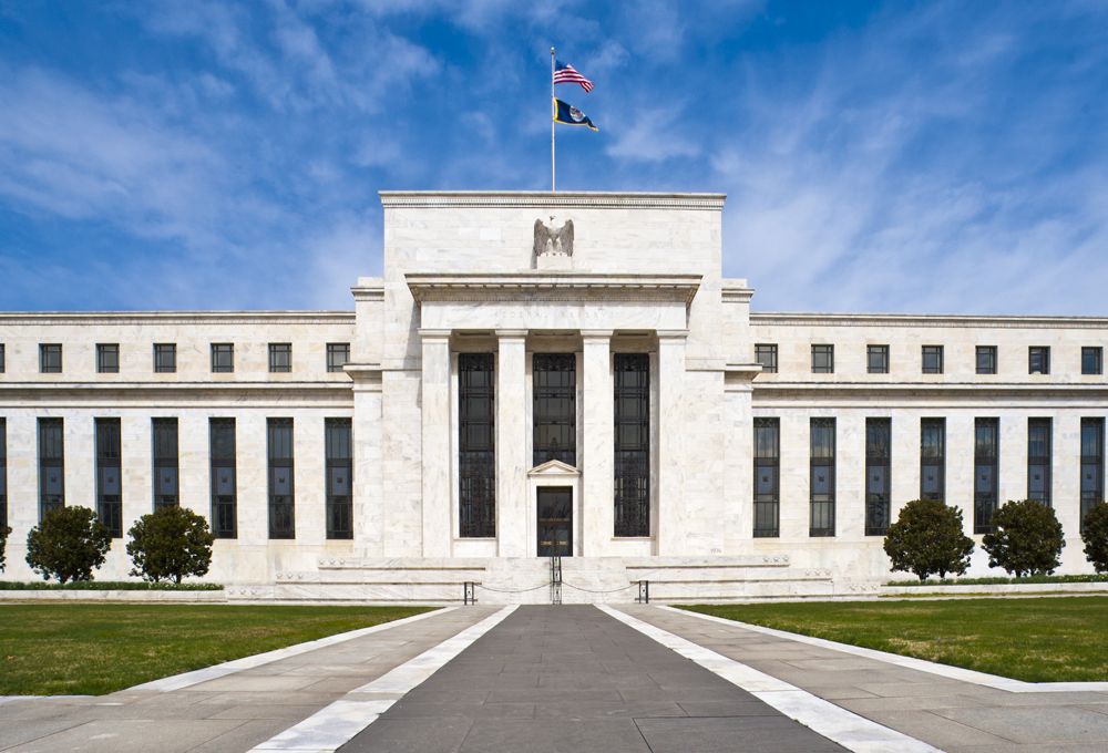 Front view of the Federal Reserve’s headquarters building, an imposing marble structure with columns and a flying American flag.