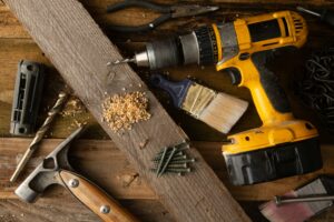 Various construction tools including a drill, hammer, screws, paintbrushes, and wood shavings on wooden boards