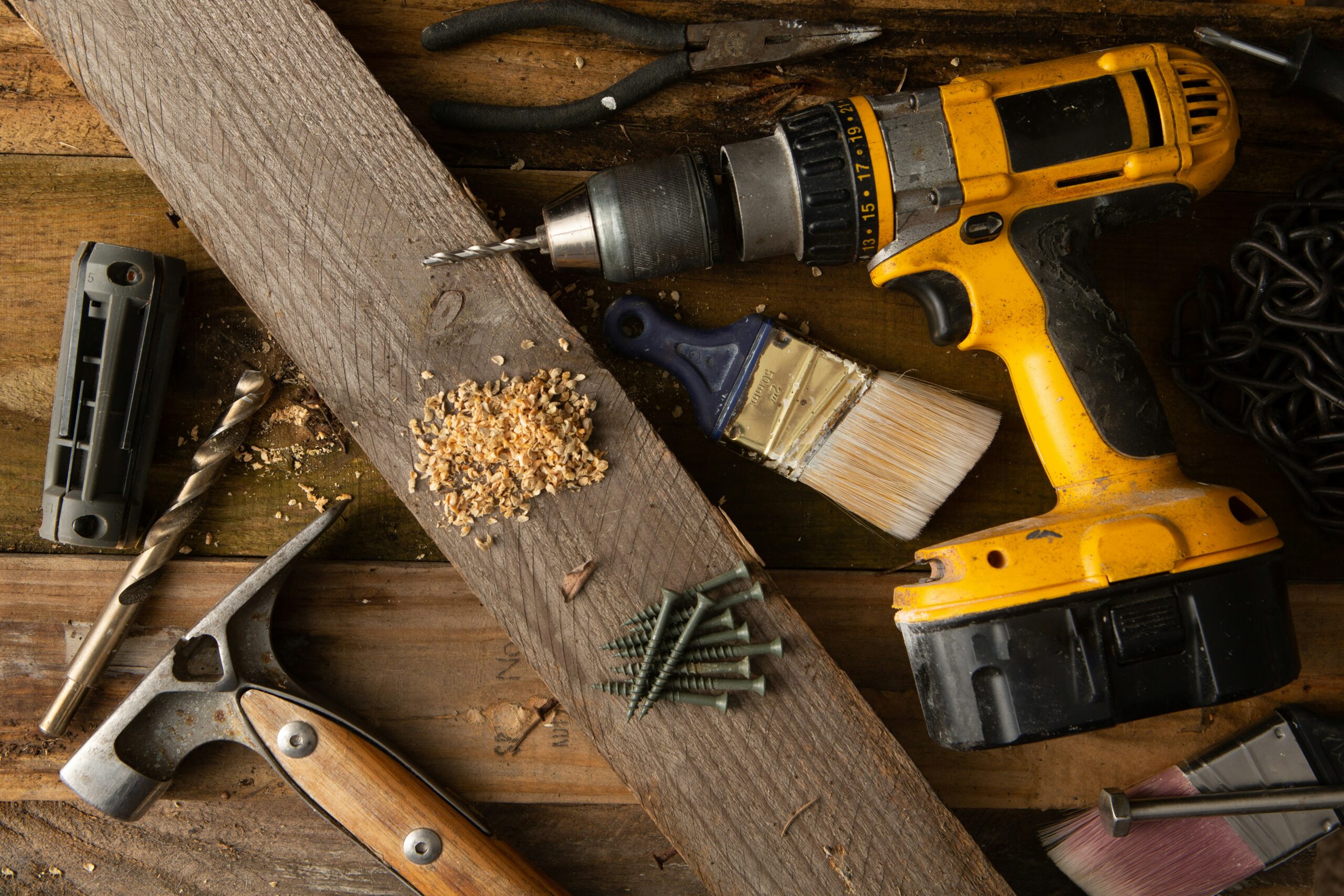 Various construction tools including a drill, hammer, screws, paintbrushes, and wood shavings on wooden boards