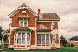 Vintage two-story Victorian-style house with faded paint, intricate detailing, and large windows on an overcast day.