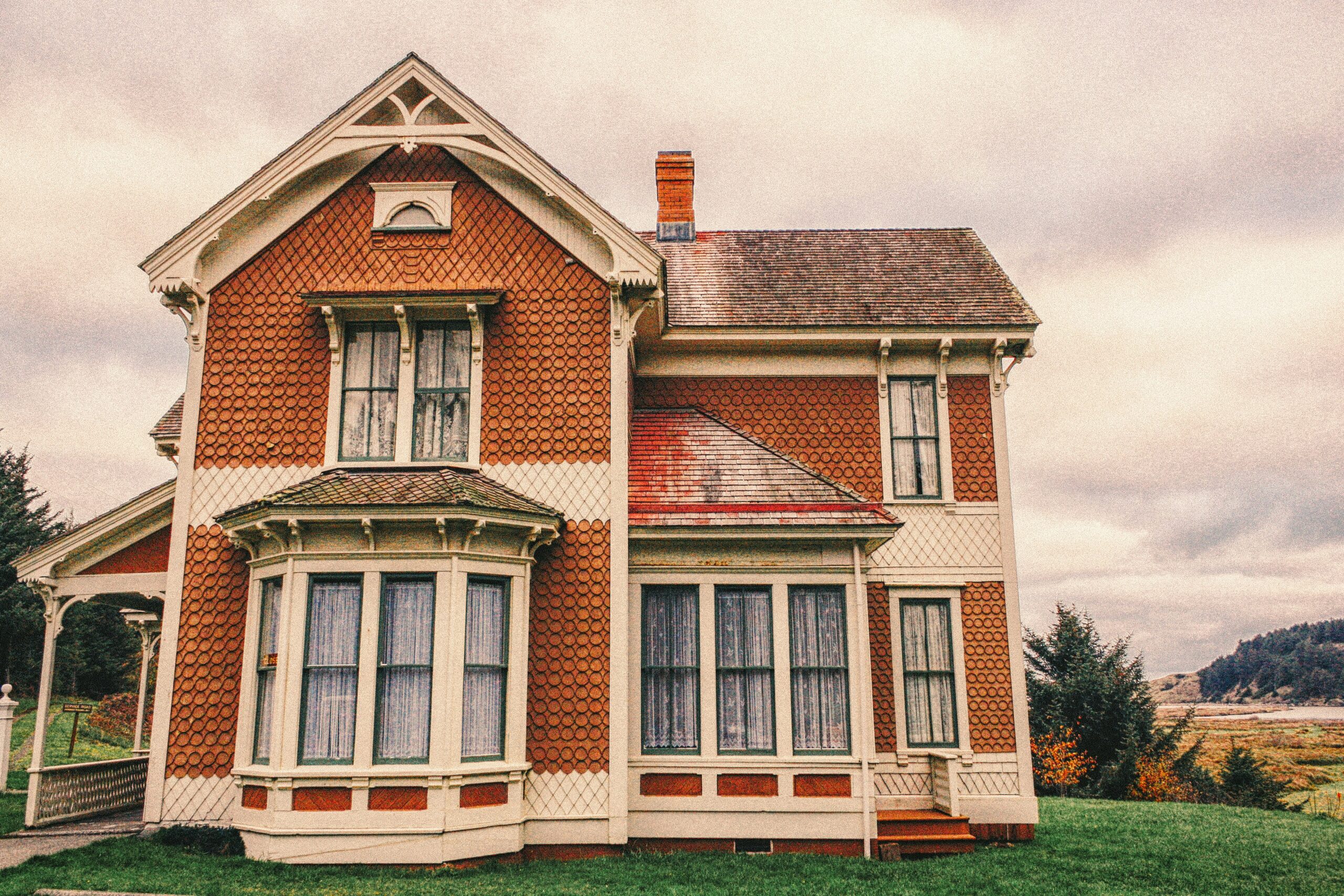 Vintage two-story Victorian-style house with faded paint, intricate detailing, and large windows on an overcast day.