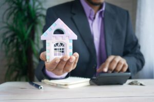 A real estate investor holding a small house model in one hand while using a calculator with the other, wearing a professional suit.