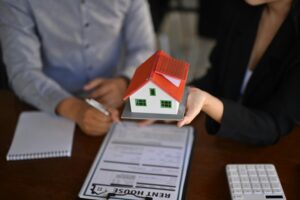 Real estate agent holding a small model house while a client fills out paperwork.